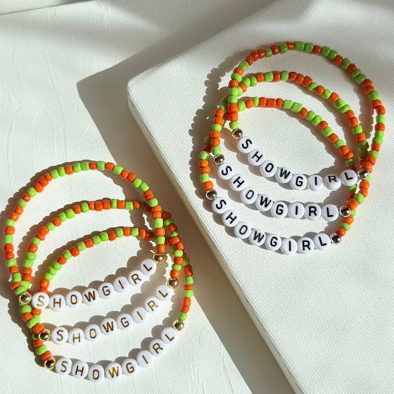 May include: Three beaded bracelets with the word "SHOWGIRL" spelled out in white letter beads. The bracelets feature a combination of orange, green, and gold beads. The bracelets are displayed on a white surface.