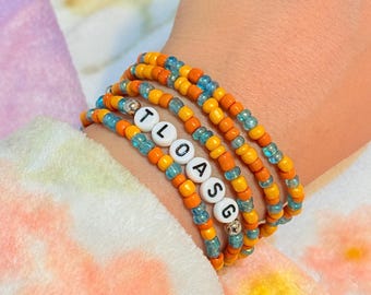 Könnte beinhalten: Ein Stapel aus vier Perlenarmbändern mit abwechselnden orangefarbenen und durchsichtigen blauen Glasperlen. Ein Armband enthält weiße runde Perlen mit den schwarzen Buchstaben T, L, O, A, S und G sowie zwei kleine silberfarbene Metallperlen.