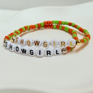 May include: Two beaded bracelets. The top bracelet has orange and green beads. The bottom bracelet has white beads with the letters "SHOWGIRL" spelled out. The top bracelet also has the words "SHOWGIRL" spelled out. The bracelets are on a white surface.
