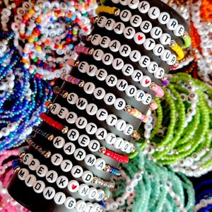 May include: A close-up of a stack of colorful beaded bracelets with white letter beads spelling out the names of Taylor Swift albums. The bracelets are arranged on a black stand. The albums include Folklore, Debut, Eras Tour, Fearless, Lover, Evermore, 1989, Reputation, Red, Karma, Speak Now, and Midnights.