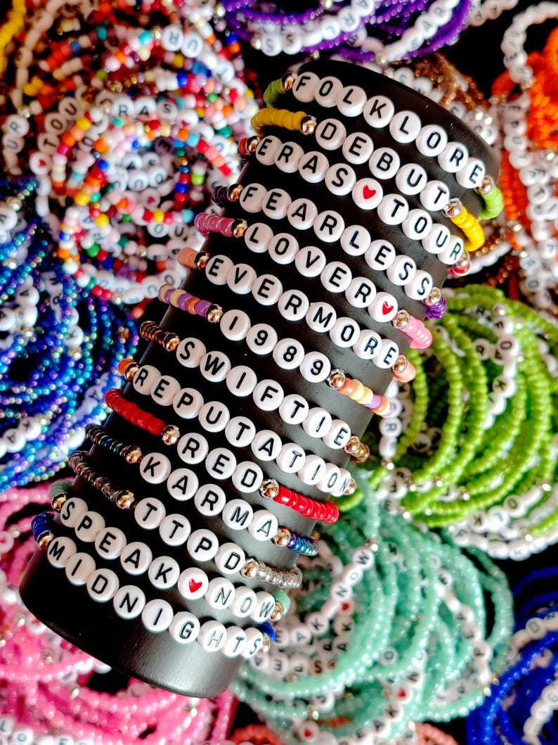 May include: A close-up of a stack of colorful beaded bracelets with white letter beads spelling out the names of Taylor Swift albums. The bracelets are arranged on a black stand. The albums include Folklore, Debut, Eras Tour, Fearless, Lover, Evermore, 1989, Reputation, Red, Karma, Speak Now, and Midnights.