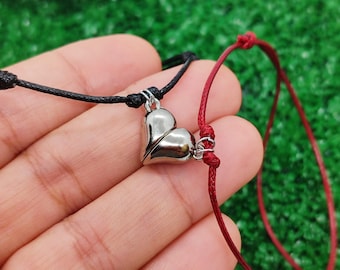 May include: Two friendship bracelets with a silver heart charm. One bracelet is red and the other is black.