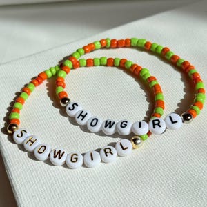 May include: Two beaded bracelets. One bracelet is orange and green, and the other is white with gold lettering spelling "SHOWGIRL". The bracelets are made of small, round beads and are likely elastic.