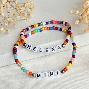 May include: Two circular bracelets made of small, multicolored glass beads. One bracelet features white letter beads spelling HELENA, and the other features white letter beads spelling MIMI. Both bracelets include small metallic silver spacer beads.