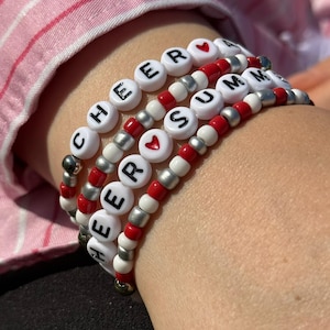 Könnte beinhalten: Ein Set aus drei Perlenarmbändern mit weißen runden Perlen, die mit schwarzen Buchstaben die Wörter CHEER und SUMMER bilden, durchsetzt mit roten Herzperlen. Die Armbänder bestehen aus abwechselnd angeordneten weißen, roten und silbernen Perlen.