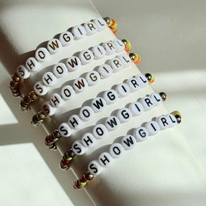 May include: Six white beaded bracelets spelling "SHOWGIRL" in gold lettering. The bracelets have colorful accents of gold, orange, green, and red. The bracelets are displayed on a white surface.
