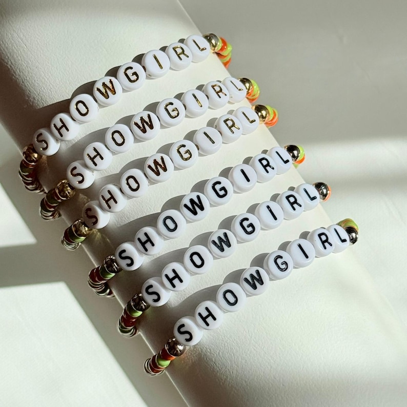 May include: Six white beaded bracelets spelling "SHOWGIRL" in gold lettering. The bracelets have colorful accents of gold, orange, green, and red. The bracelets are displayed on a white surface.
