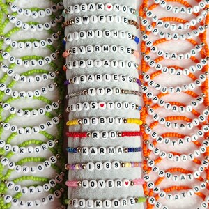 May include: A collection of beaded bracelets in various colors, each spelling out words and phrases like "FOLKLORE," "REPUTATION," and song titles. The bracelets feature white letter beads and colorful accent beads.
