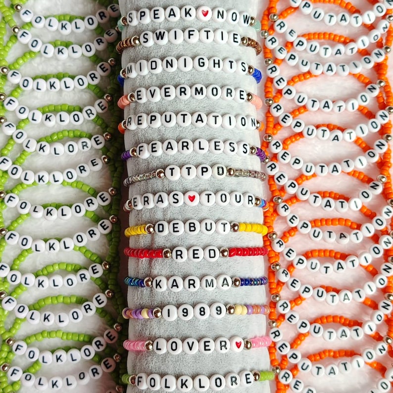 May include: A collection of beaded bracelets in various colors, each spelling out words and phrases like "FOLKLORE," "REPUTATION," and song titles. The bracelets feature white letter beads and colorful accent beads.