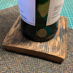 Pode incluir: Um apoio de copo de madeira feito de um barril de u&iacute;sque, com um acabamento castanho escuro. O apoio de copo tem um recorte circular no centro para segurar uma garrafa. A garrafa &eacute; de vidro verde e tem um r&oacute;tulo com o texto "The Single Malt Whisky".