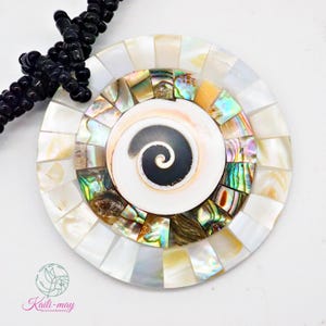 May include: A circular pendant featuring a central spiral shell piece surrounded by concentric rings of iridescent abalone shell segments and white mother-of-pearl tiles. The pendant is attached to a strand of small, round, glossy black beads.
