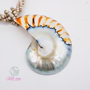 May include: A nautilus shell pendant featuring a polished, iridescent surface with orange and black striped patterns, suspended from a multi-strand necklace made of small, light beige cylindrical beads. The logo Kaili-may is visible.