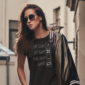 Peut inclure: Une femme portant un t-shirt noir avec le texte "Eat Clean, Lift Heavy, Code Daily, Repeat!" et un pantalon à carreaux noir et blanc avec des broderies florales. Elle porte des lunettes de soleil et une veste en cuir noire.