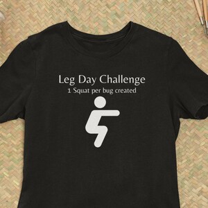 Op de afbeelding: Zwart T-shirt met de tekst "Leg Day Challenge" en een witte afbeelding van een persoon in squatpositie. De tekst eronder luidt "1 Squat per bug created". Het T-shirt ligt op een geweven mat.