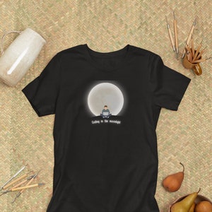 Peut inclure: T-shirt noir avec un graphique d'une personne assise devant une grande lune, codant sur un ordinateur portable. Le texte "Coding in the moonlight" est sous l'image.
