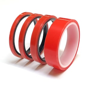 May include: A collection of red double-sided tape rolls on a white backdrop. The tape features a vibrant red outer layer, a white inner layer, and a black band. The rolls are curved, highlighting the product's design and structure.