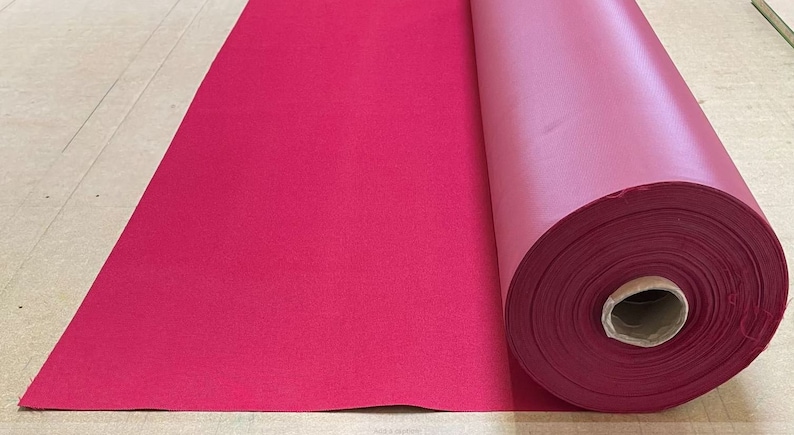 May include: A roll of deep red fabric, unfurled on a light brown surface. The fabric is smooth and has a slight sheen.