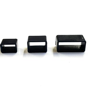 May include: Three black rectangular rubber loops of different sizes are displayed against a white background. Each loop has a hollow, rectangular centre. The smallest loop is square, while the largest is more elongated in shape.