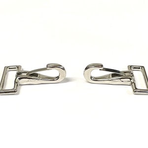 May include: Two silver metal snap hooks with a rectangular loop on each side. The hooks are closed and ready to be attached to a strap or other item.