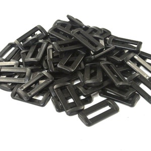 May include: A collection of black plastic rectangular buckles, possibly for straps or belts, on a white backdrop. The buckles feature a straightforward design with a rectangular aperture in the middle.