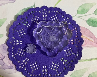 May include: A clear plastic spider web and spider stamp set on a purple doily. The spider web stamp is shaped like a heart.