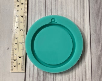 May include: A teal-colored, circular silicone mold with a raised outer edge and a small loop at the top. A ruler is placed next to the mold, showing measurements in inches. The mold is designed for crafting.