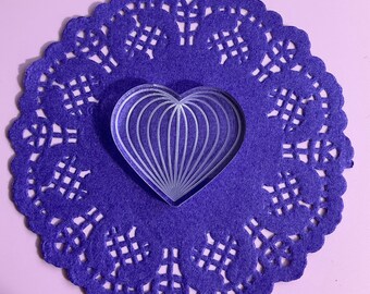 May include: A clear plastic heart-shaped stamp with a decorative design on a purple doily.