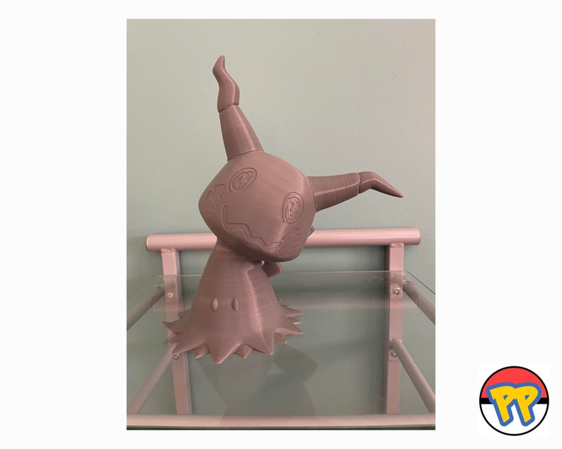 May include: A grey 3D-printed Mimikyu figure, a ghost and fairy-type Pok&eacute;mon. The figure has a rounded body, two arms, and a tall, curved horn. The figure is displayed on a glass shelf with a light blue wall in the background.