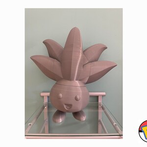 Op de afbeelding: Een grijs 3D-geprint beeldje van een Pok&eacute;mon-personage met een groot, bladerrijk hoofd en een glimlachend gezicht. Het beeldje staat op een glazen plank.