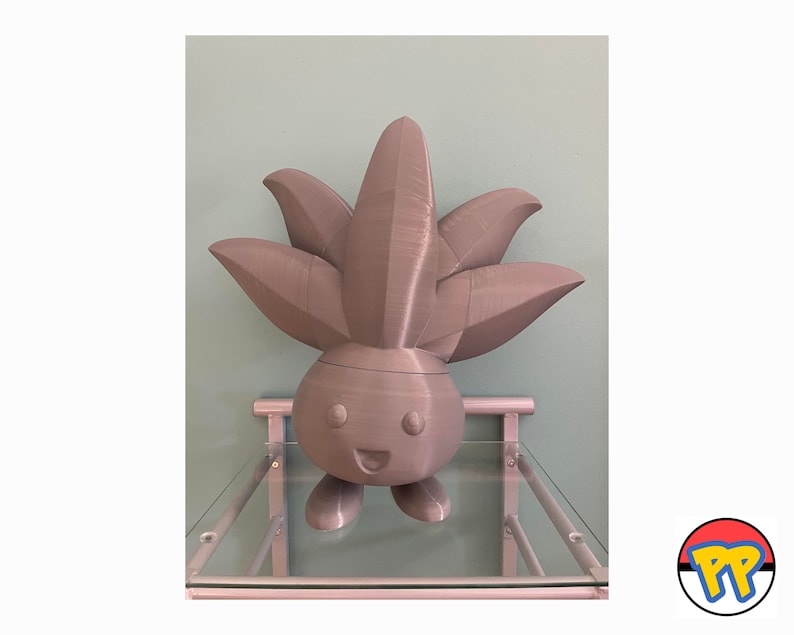 Op de afbeelding: Een grijs 3D-geprint beeldje van een Pok&eacute;mon-personage met een groot, bladerrijk hoofd en een glimlachend gezicht. Het beeldje staat op een glazen plank.