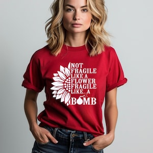 May include: A red t-shirt featuring the white text "NOT FRAGILE LIKE A FLOWER FRAGILE LIKE A BOMB". The design includes a white sunflower and a bomb graphic. The shirt is being worn.