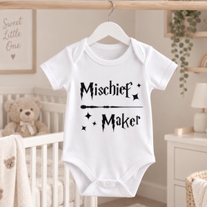 May include: A white short-sleeved baby bodysuit featuring black text that reads Mischief Maker. The design includes a horizontal wand graphic between the two words, with lightning bolt accents on the letters M and star-shaped sparkles.