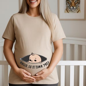 May include: A beige maternity t-shirt with a cartoon baby design peeking out of a zipper. The text "Hello, is it time yet?" is printed below the baby. The woman is smiling and holding her belly.