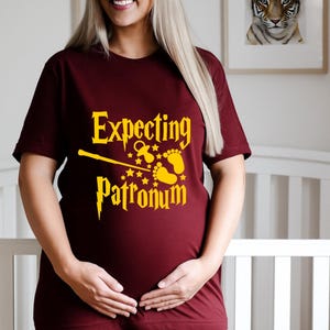 May include: A maroon t-shirt with the words "Expecting Patronum" in yellow, featuring a wand, baby feet, stars, and a pacifier graphic. The short-sleeved shirt is worn by a person. The background includes a cot and a framed tiger print.