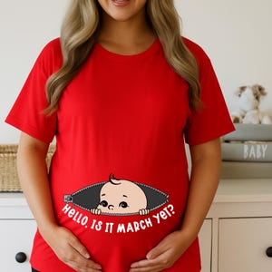May include: A red t-shirt featuring a baby graphic emerging from a zipper design. The text "Hello, Is it March yet?" is printed below. The shirt has short sleeves and is a maternity top.