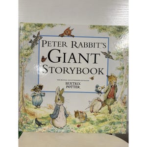 Puede incluir: Un libro de tapa dura titulado "Peter Rabbit's Giant Storybook" con ilustraciones de personajes de Beatrix Potter. La portada presenta un borde azul e imágenes de conejos, patos y otros animales en un jardín.