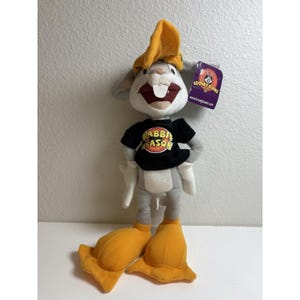 May include: A plush Bugs Bunny toy wearing a yellow hat and a black t-shirt with the text RABBIT SEASON printed in yellow and red. The character has grey fur, white gloves, and large yellow feet. A purple Looney Tunes tag is attached to the ear.