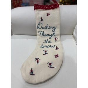 May include: Cream-colored Christmas stocking with a red polka-dot cuff and a red hanging loop. The words "Dashing Through the Snow" are embroidered in teal, along with skiers and sleds. Small sequins are scattered across the stocking.