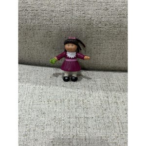 May include: A small plastic doll with brown hair, wearing a purple dress with a white collar and cuffs. The doll has black shoes and is holding a small green object. The doll's face is painted with a smiling expression.