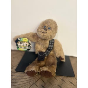 Op de afbeelding: Een bruine Chewbacca-knuffelpop uit de Star Wars-franchise. De pop heeft een zwarte bandolier en een klein zwart zakje. Blauwe ogen en een label met "Star Wars".