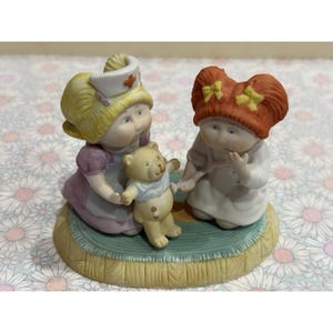 May include: A ceramic figurine depicting two children and a teddy bear. One child wears a nurse's cap and a purple dress, while the other has red hair and a white dress. The teddy bear has a blue shirt. The figurine is on a round base.