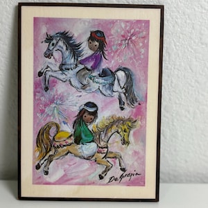 May include: A rectangular wall art print featuring a DeGrazia illustration of two figures riding horses against a pink background with starburst patterns. The top figure wears purple, the bottom figure wears green, and the print has a dark wood frame.