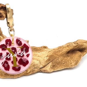 Könnte beinhalten: Ein runder rosa Schlüsselanhänger mit einem goldenen Buchstaben Y und dem Wort "Yuna" in weißer Kursivschrift. Der Schlüsselanhänger ist mit roten Blütenblättern und goldenem Glitzer verziert.