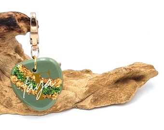 May include: A green and gold resin keychain with the letter "T" and the name "Taya" written in white. The keychain is attached to a gold-colored metal ring.