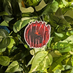Op de afbeelding: Een sticker met een cirkelvormig ontwerp, een rode rand en een centraal beeld van een persoon. De sticker heeft een witte rand en is omgeven door groene bladeren. De persoon in het midden draagt donkere kleding.