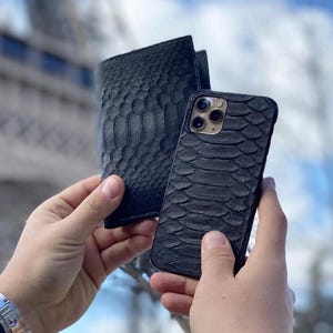 May include: Black leather wallet and phone case with a reptile skin texture. The wallet is folded, and the phone case is designed for a smartphone. The background features a blurred view of a tall structure and a blue sky.