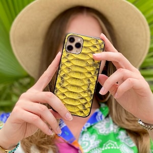May include: A yellow snakeskin-effect phone case with a black frame. The phone case is held up by a person wearing a colourful patterned shirt and a straw hat. Green palm leaves are in the background.