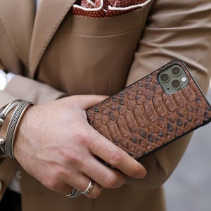 May include: A tan suit with a white shirt and a red polka dot pocket square. A brown textured phone case is held. Silver bracelets and a watch are also visible.