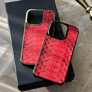 Peut inclure: Deux coques de téléphone rouges avec une texture peau de reptile. L'une a un cadre doré, l'autre un cadre noir. Les deux coques reposent sur une boîte noire.