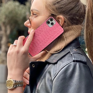 May include: A pink textured phone case with a reptile skin pattern. The person is wearing a black leather jacket, gold-rimmed glasses, and a gold watch with a silver band. The phone is held up to the ear.